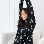 Woman stretching wearing black long sleeve button up pajamas in a magnolia print against a white wooden wall