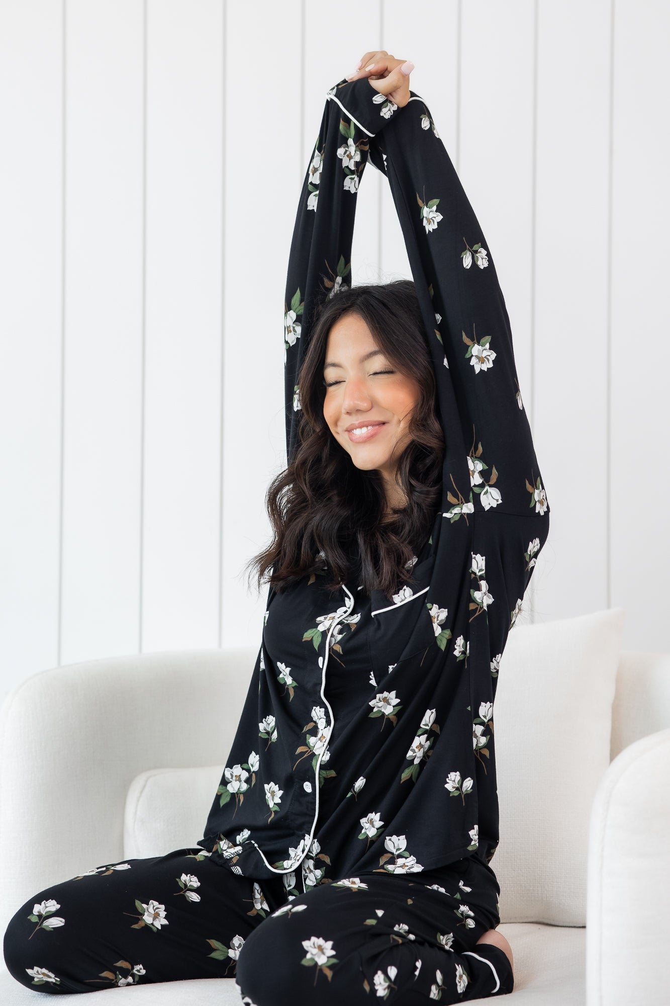 Woman stretching wearing black long sleeve button up pajamas in a magnolia print against a white wooden wall