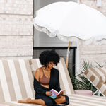 Woman wearing black robe and dark brown bralette reading a book on a striped lounge chair under an umbrella.