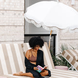 Woman wearing black robe and dark brown bralette reading a book on a striped lounge chair under an umbrella.