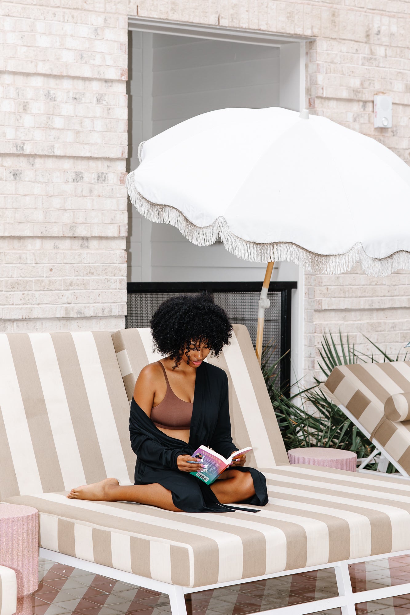 Woman wearing black robe and dark brown bralette reading a book on a striped lounge chair under an umbrella.