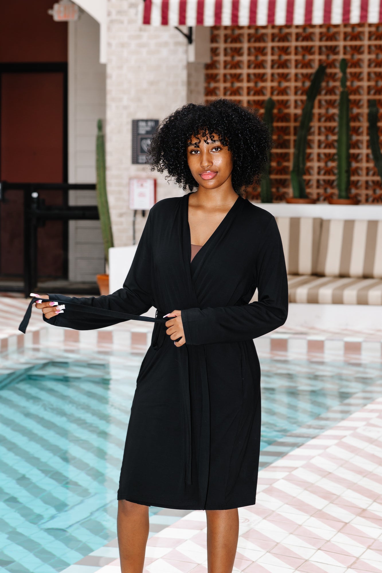 Woman in a black robe standing by a poolside with a striped awning above.