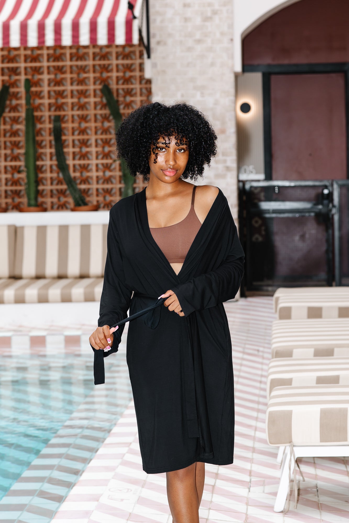 Woman in a black robe standing by a poolside with a striped umbrella in the background