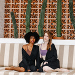 Two women in black robes sitting on a striped couch with cacti in the background
