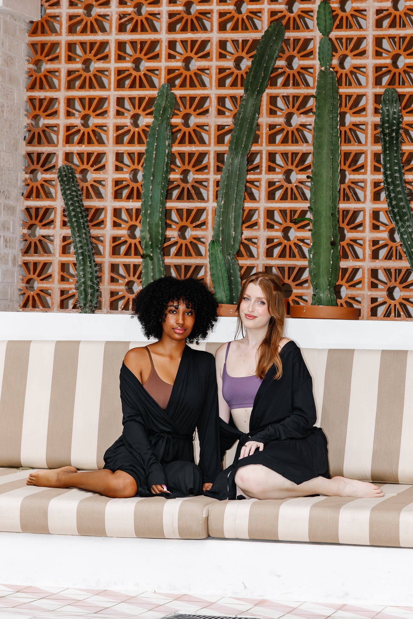 Two women in black robes sitting on a striped couch with cacti in the background