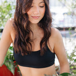 Woman wearing a black bralette and beige pant with plants in the background