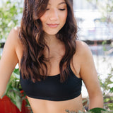 Woman wearing a black bralette and beige pant with plants in the background