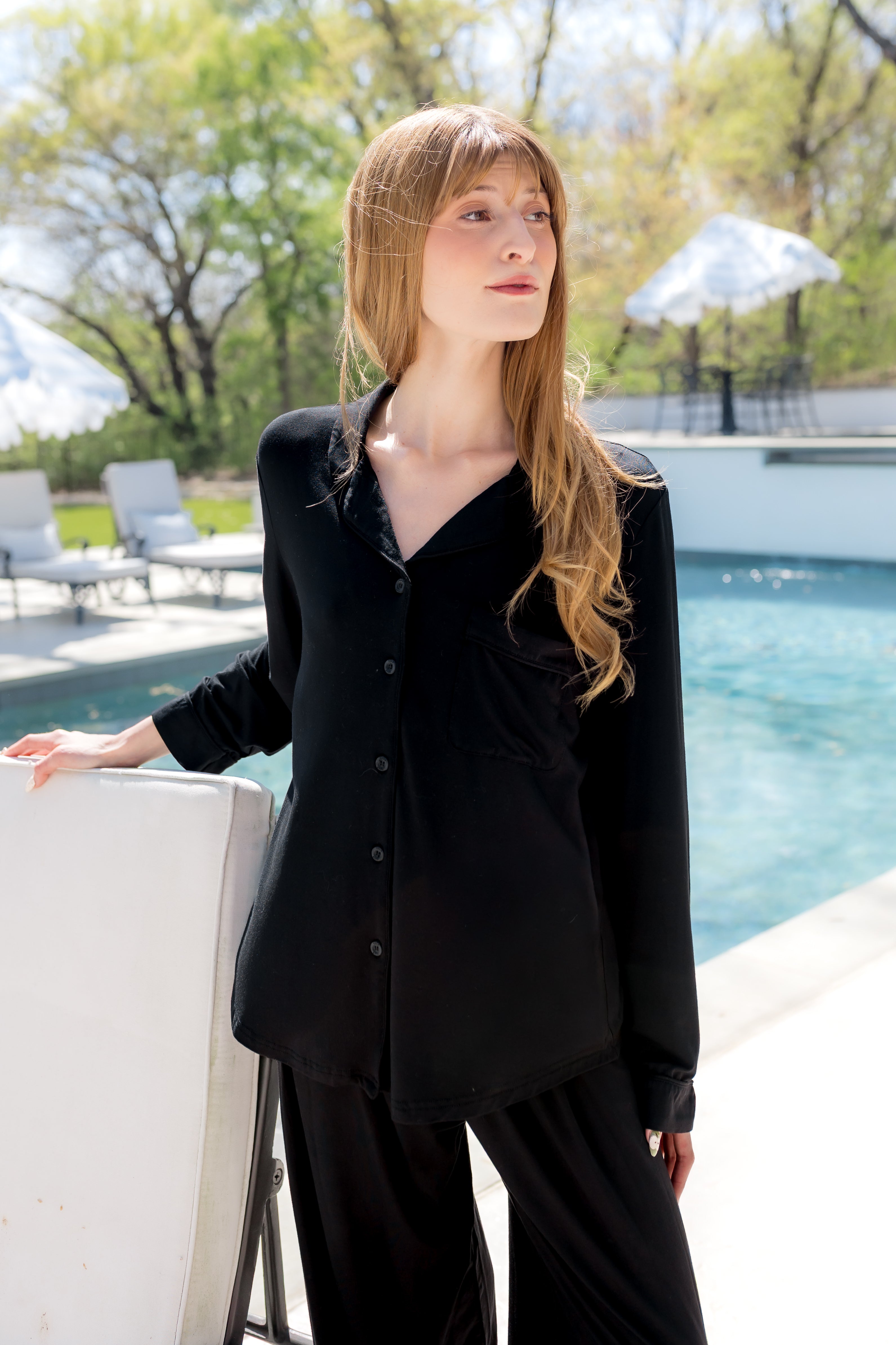 Woman in a black pajama set standing by a pool with trees and umbrellas in the background