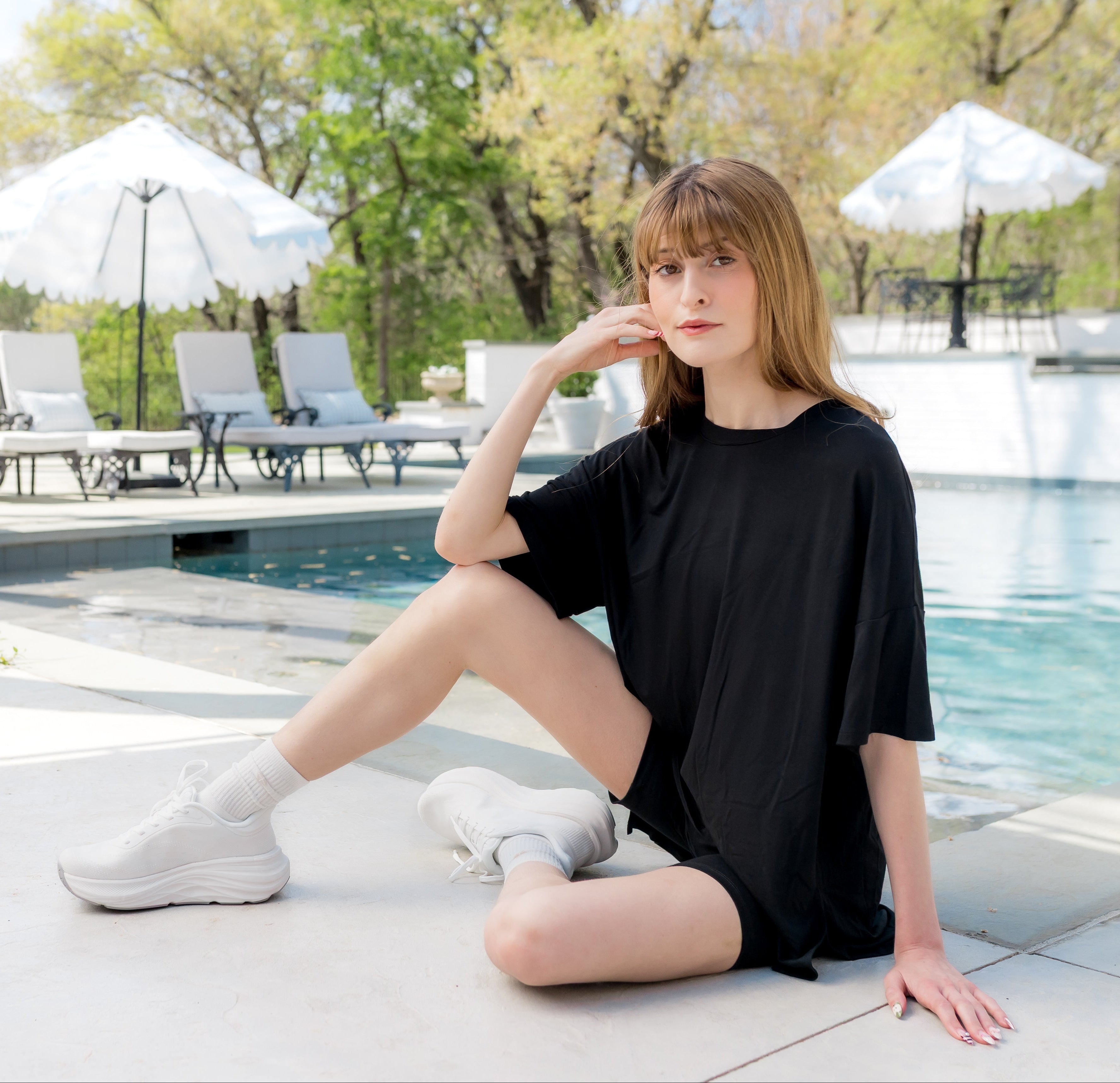 Woman in a black outfit sitting by a pool with trees and chairs in the background