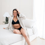 Woman in black intimates set and oat robe sitting on a bed.