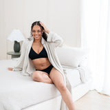 Woman in black intimates set and oat robe sitting on a bed.