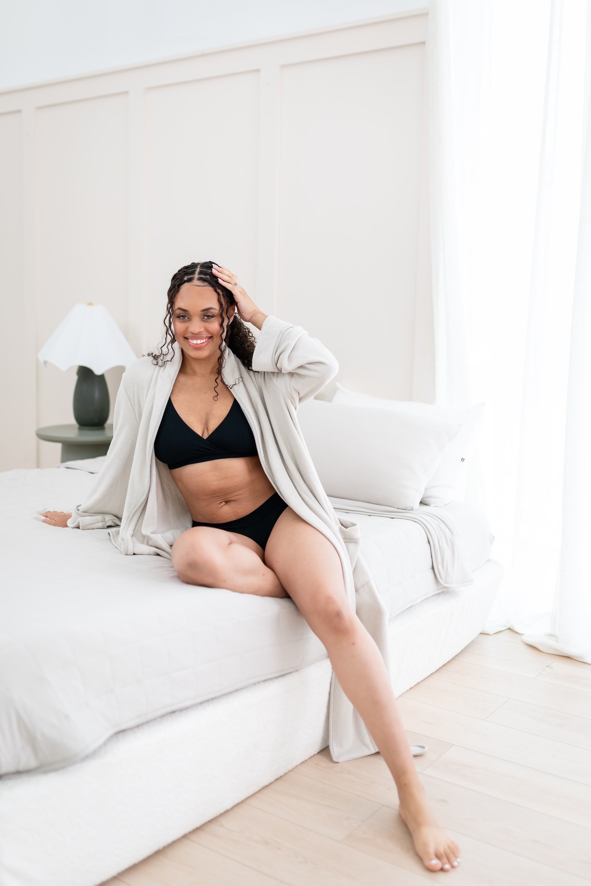 Woman in black intimates set and oat robe sitting on a bed.