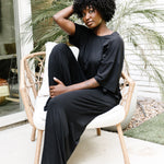 Woman sitting on a wicker chair on white cushion with her eyes closed wearing the Women's Wide Leg Pant in Midnight with matching women's short sleeve tee