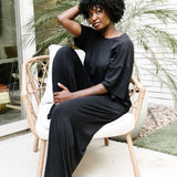 Woman sitting on a wicker chair on white cushion with her eyes closed wearing the Women's Wide Leg Pant in Midnight with matching women's short sleeve tee