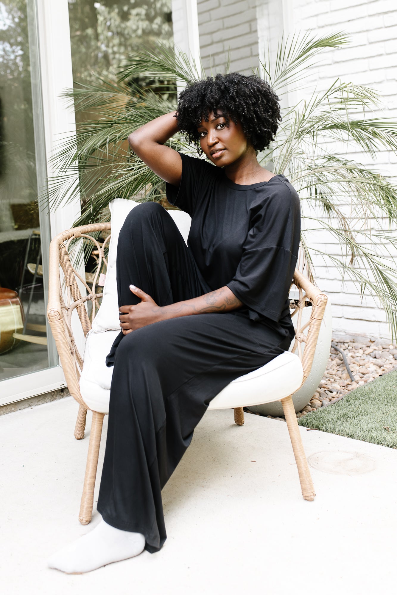Woman sitting on a wicker chair on white cushion with her eyes closed wearing the Women's Wide Leg Pant in Midnight with matching women's short sleeve tee