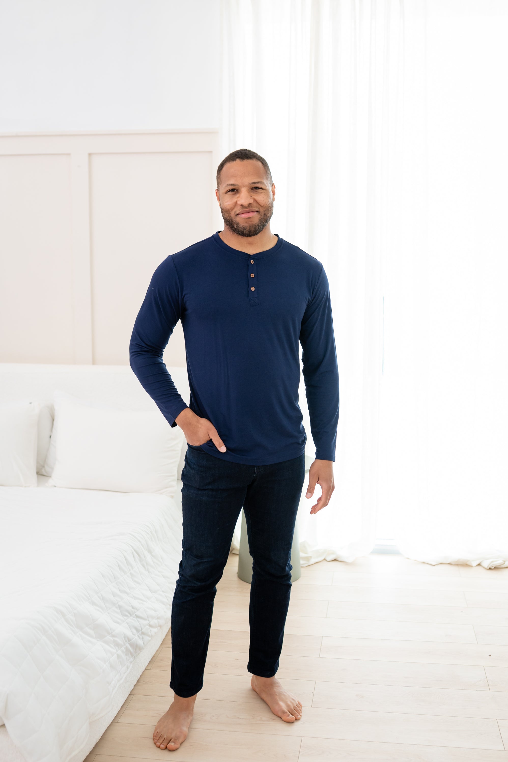 Man modeling Men's Long Sleeve Henley Top in Navy with jeans with hand in pocket with a bedroom backdrop