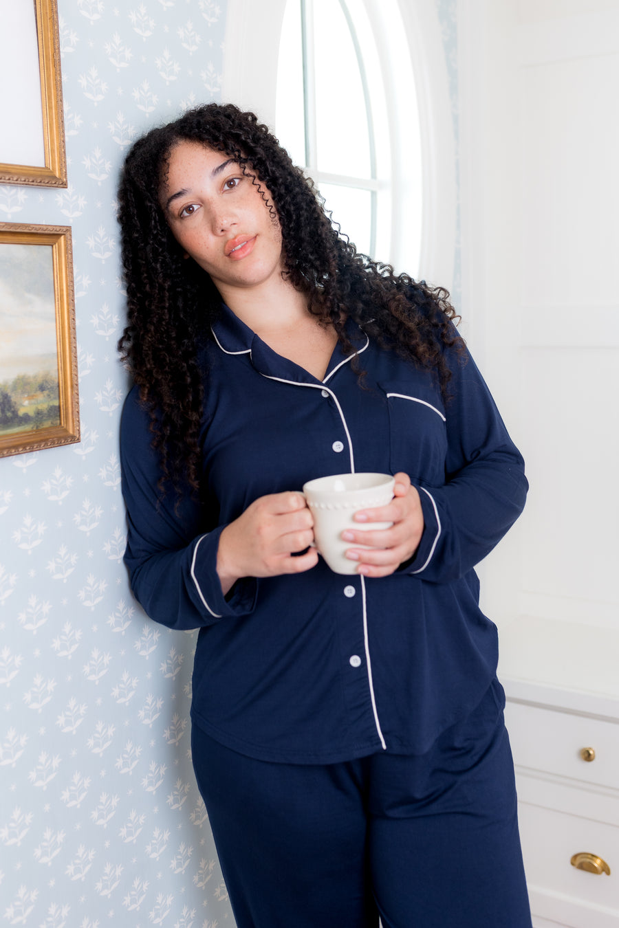 Woman in navy pajamas holding a white mug in a room with decorative wallpaper.