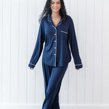 Woman modeling long sleeve button up navy pajamas with white piping against a white wooden wall