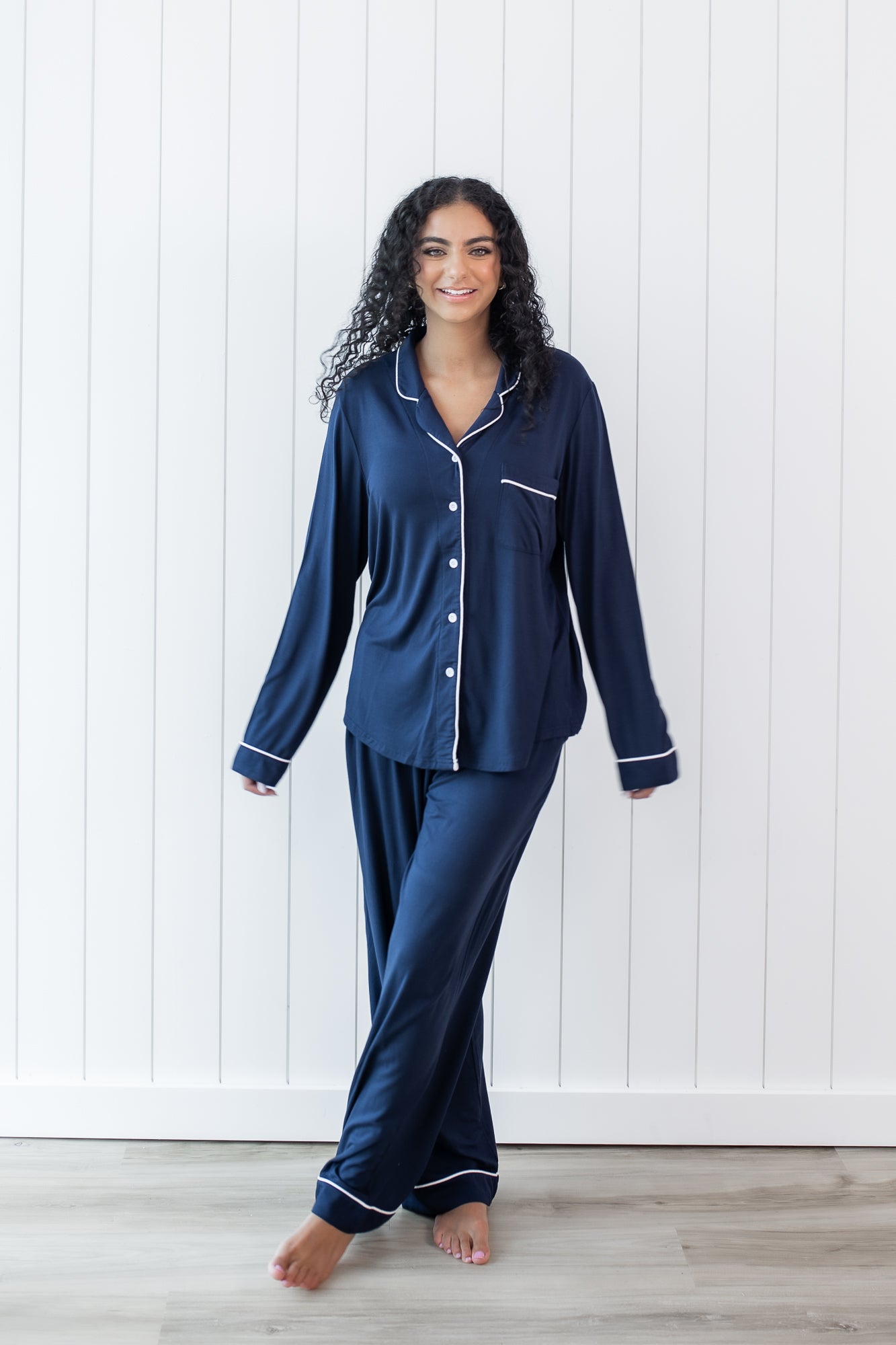 Woman modeling long sleeve button up navy pajamas with white piping against a white wooden wall