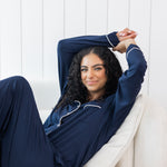 Woman modeling long sleeve button up navy pajamas with white piping sitting in cream chair
