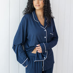 Woman modeling long sleeve button up navy pajamas with white piping against a white wooden wall showing waistband