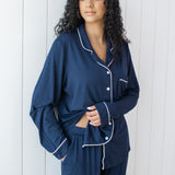 Woman modeling long sleeve button up navy pajamas with white piping against a white wooden wall showing waistband