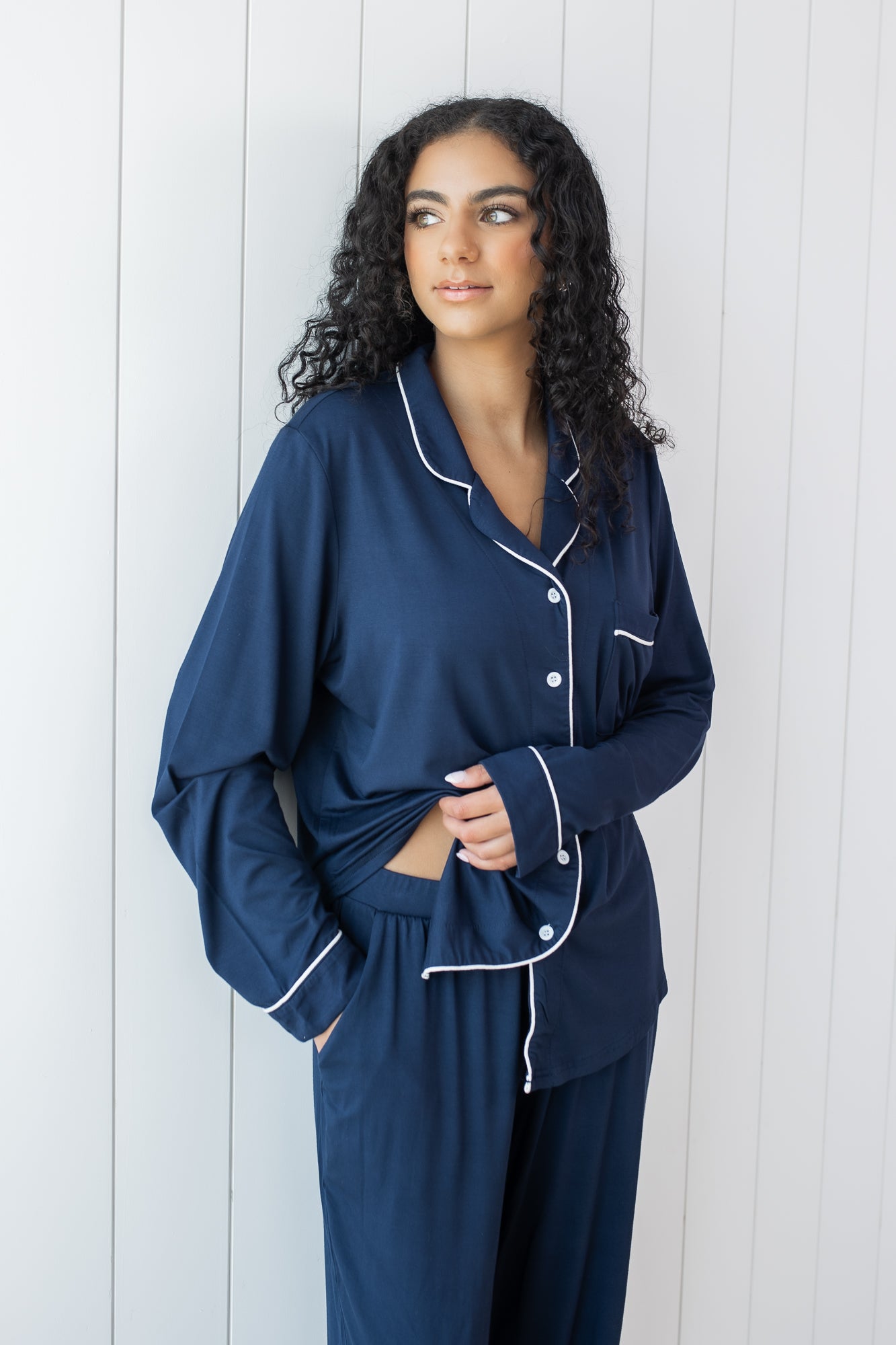 Woman modeling long sleeve button up navy pajamas with white piping against a white wooden wall showing waistband