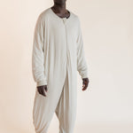 A man wearing a light beige, long-sleeve, full-body sleep sack standing against a solid, warm off-white studio background.