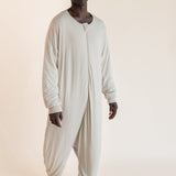 A man wearing a light beige, long-sleeve, full-body sleep sack standing against a solid, warm off-white studio background.