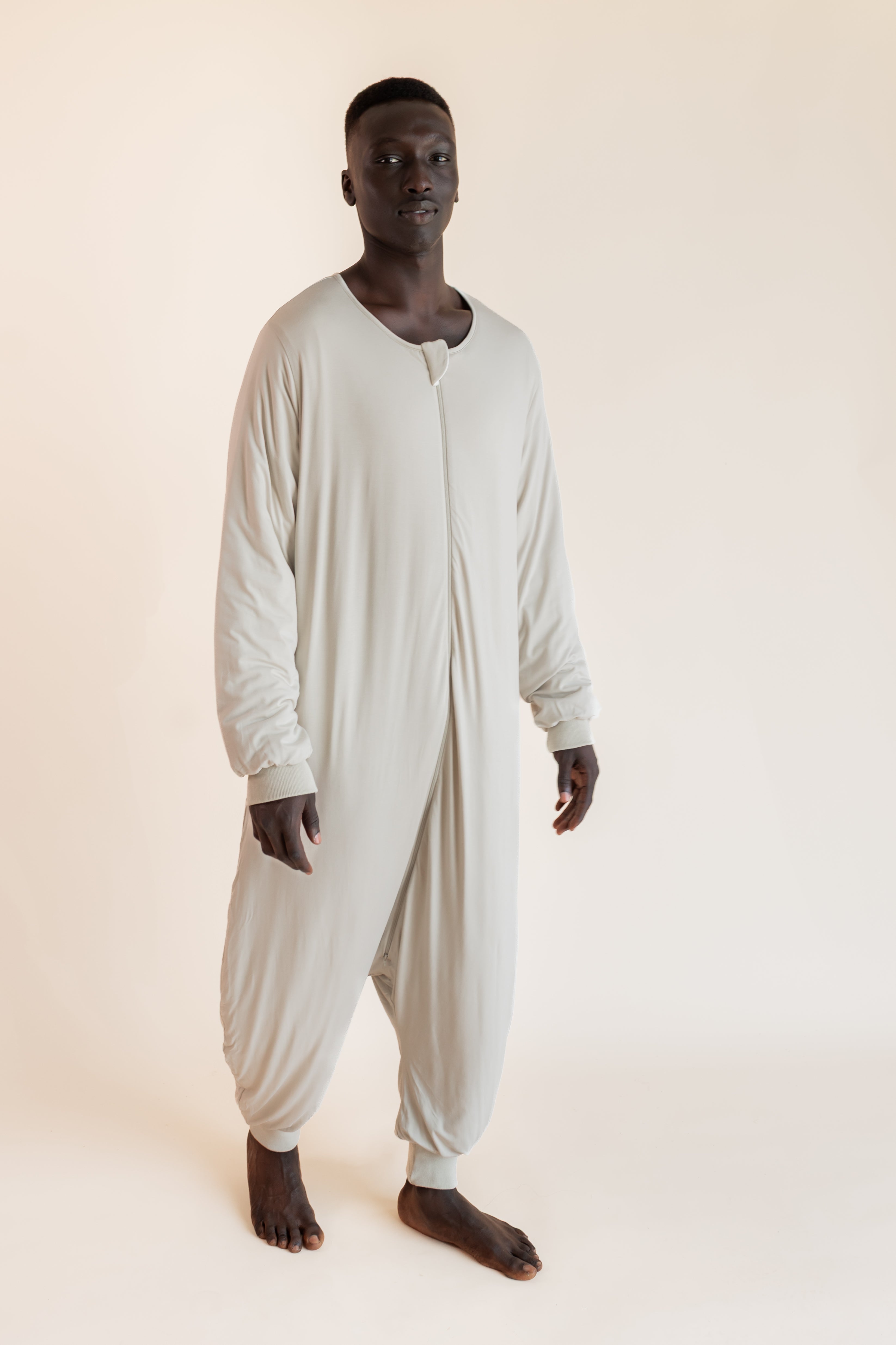 A man wearing a light beige, long-sleeve, full-body sleep sack standing against a solid, warm off-white studio background.