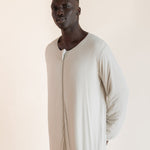 A man wearing a long-sleeved, full-length cream-colored sleep sack , standing against a solid light beige background.