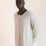 A man wearing a long-sleeved, full-length cream-colored sleep sack , standing against a solid light beige background.