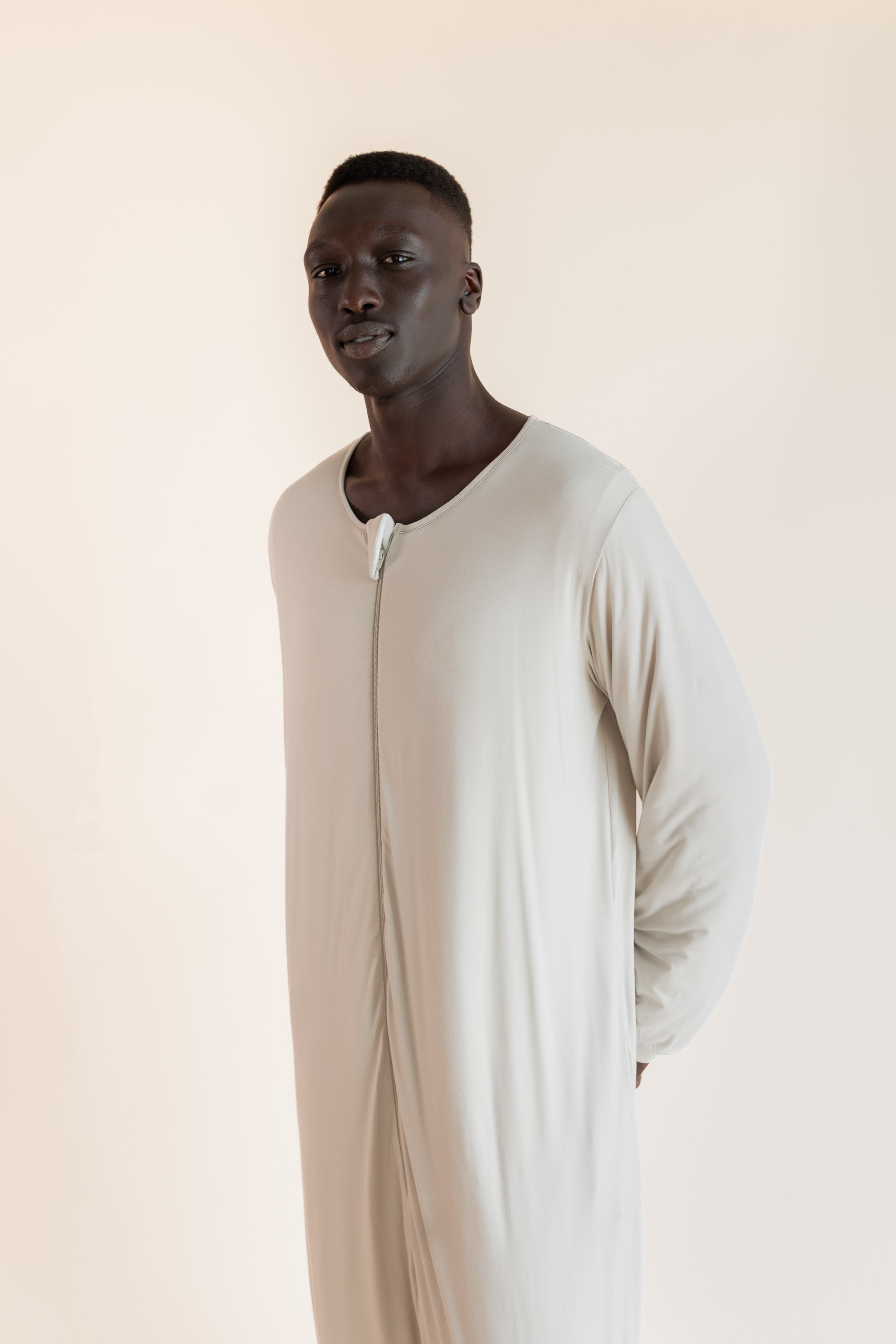 A man wearing a long-sleeved, full-length cream-colored sleep sack , standing against a solid light beige background.
