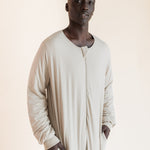 A man wearing a long-sleeved, beige sleep sack with a split neckline and side pockets, standing against a solid light beige background.