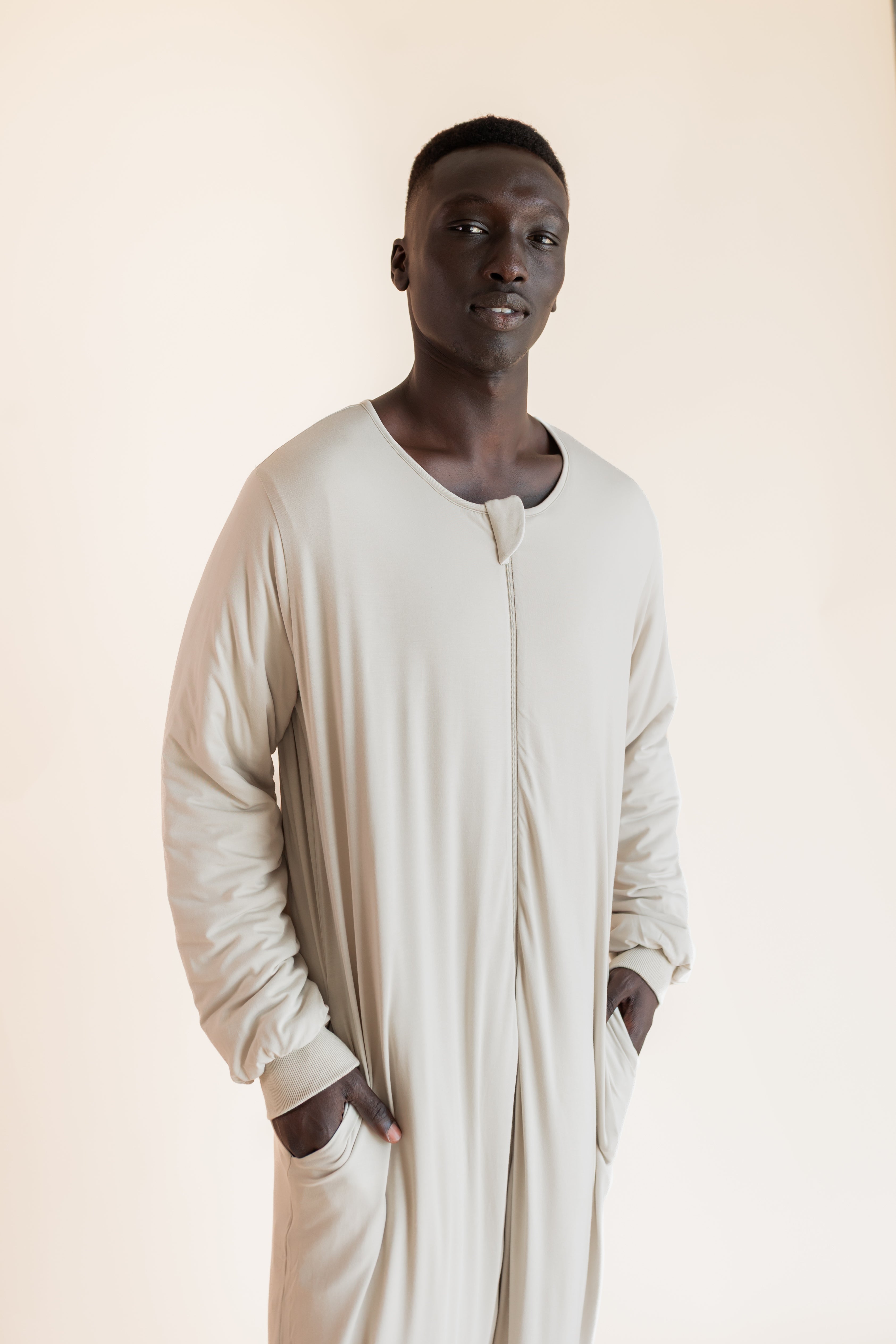A man wearing a long-sleeved, beige sleep sack with a split neckline and side pockets, standing against a solid light beige background.