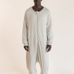 A man wearing a light beige, full-body zip-up harem sleep sack against a solid, warm off-white background.