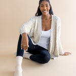 Woman sitting on a beige floor wearing a cream cardigan, black pants, and cream socks.