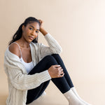 Woman wearing a beige cardigan, white top, black leggings, and cream socks sitting on a beige background