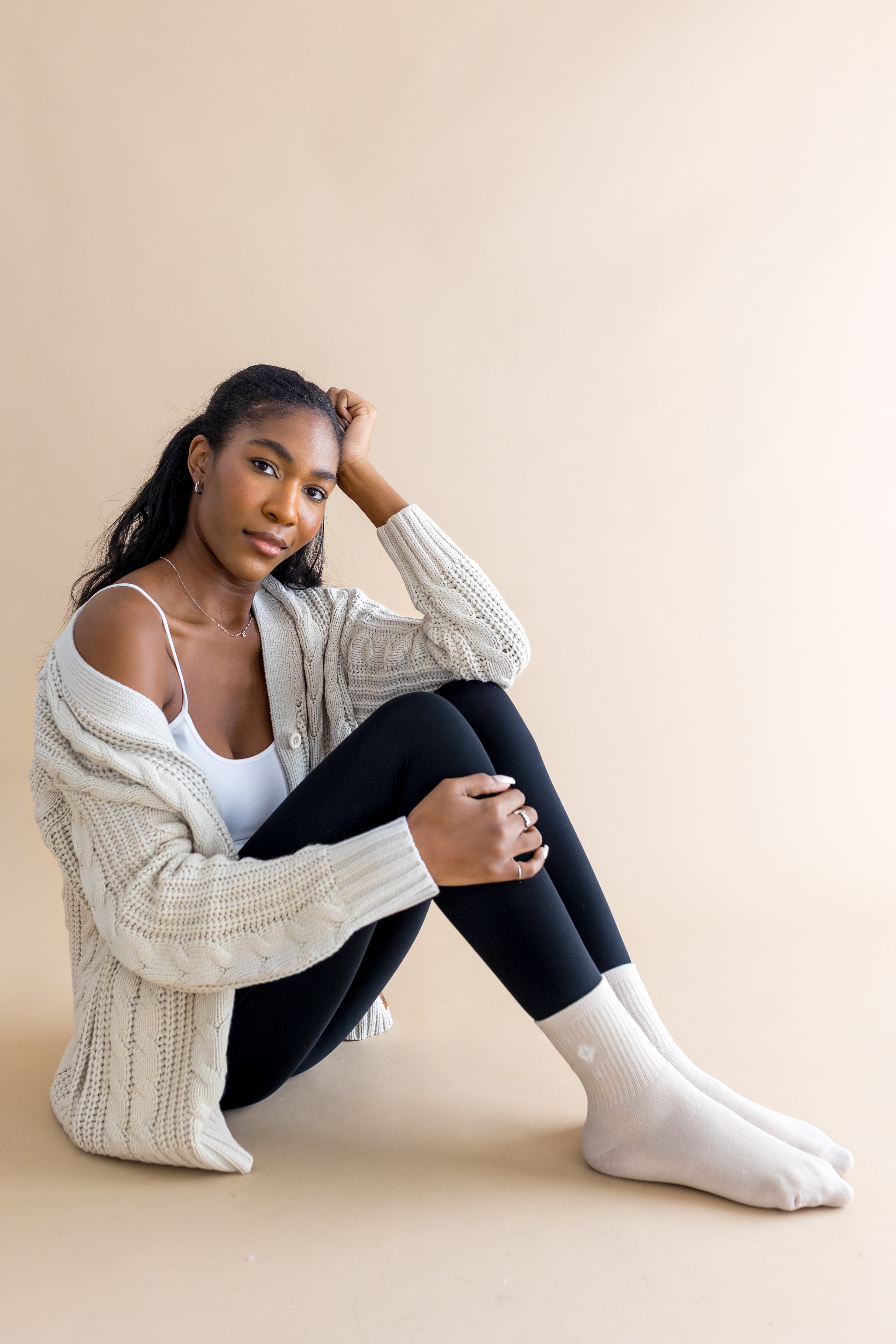 Woman wearing a beige cardigan, white top, black leggings, and cream socks sitting on a beige background