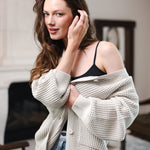 Modal posing in a living room wearing Chunky Knit Women's Waffle Cardigan in Oat over black camisole and blue jeans