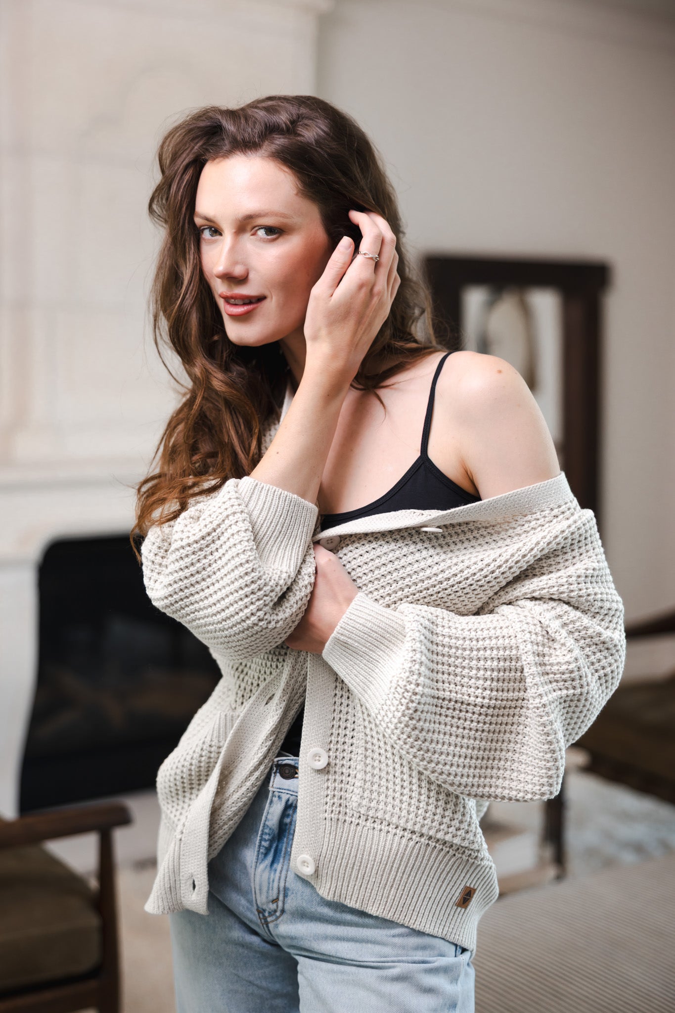 Modal posing in a living room wearing Chunky Knit Women's Waffle Cardigan in Oat over black camisole and blue jeans