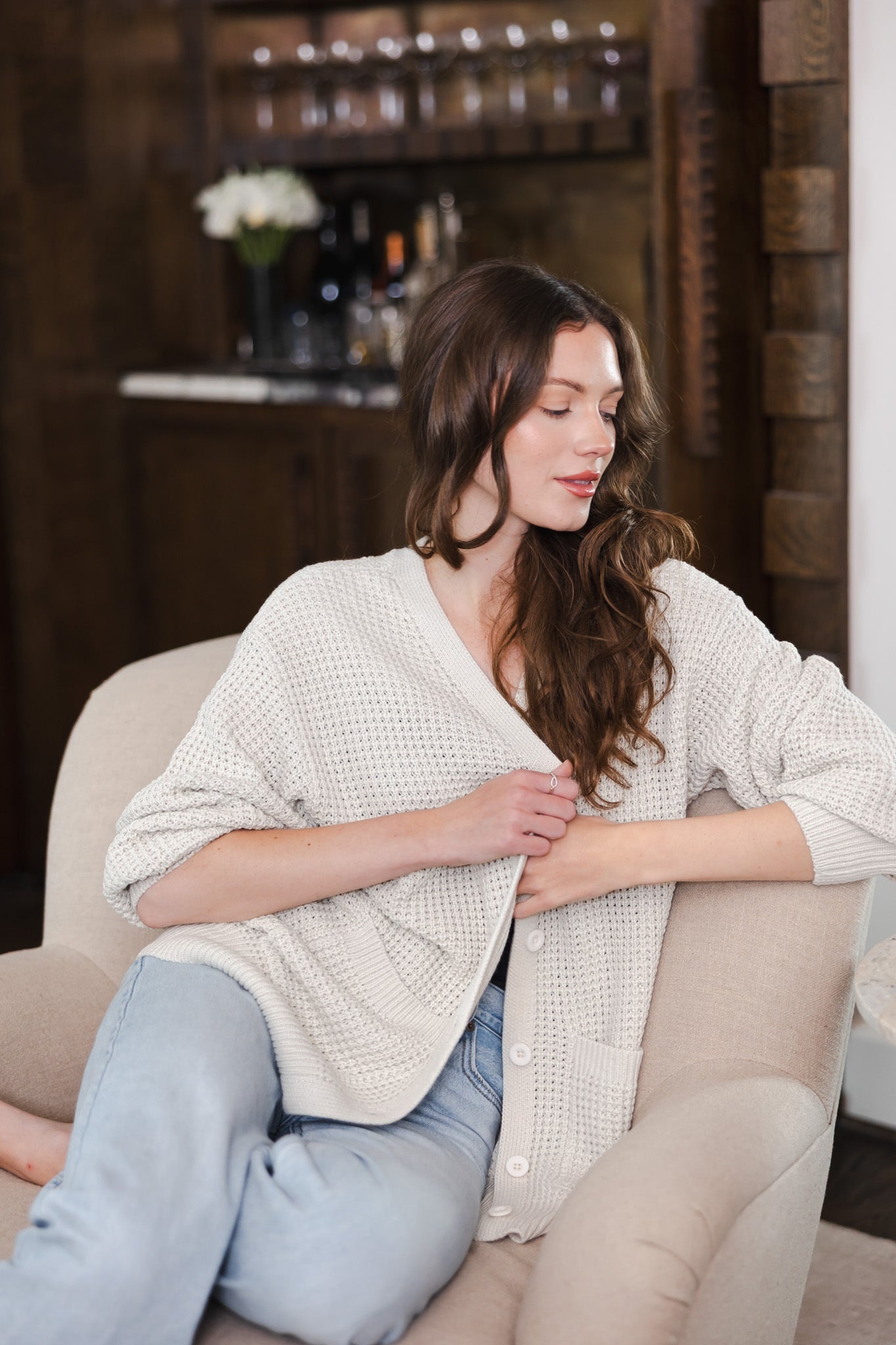 Modal sitting on white sofa in a living room wearing Chunky Knit Women's Waffle Cardigan in Oat over blue jeans
