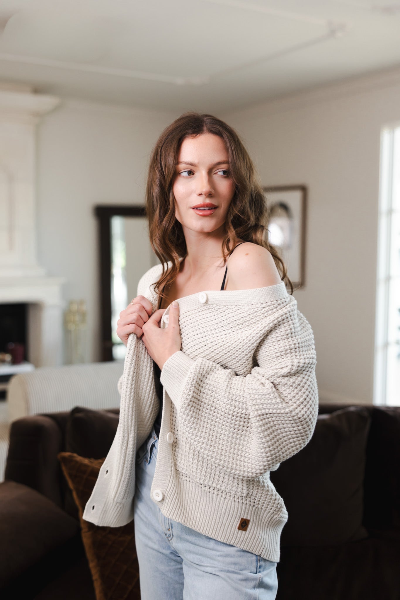 Modal standing in a living room wearing Chunky Knit Women's Waffle Cardigan in Oat over black camisole and blue jeans
