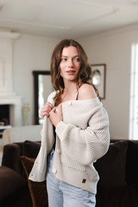 Modal standing in a living room wearing Chunky Knit Women's Waffle Cardigan in Oat over black camisole and blue jeans