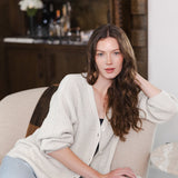Modal sitting in a living room on a sofa wearing Chunky Knit Women's Waffle Cardigan in Oat over black camisole and blue jeans