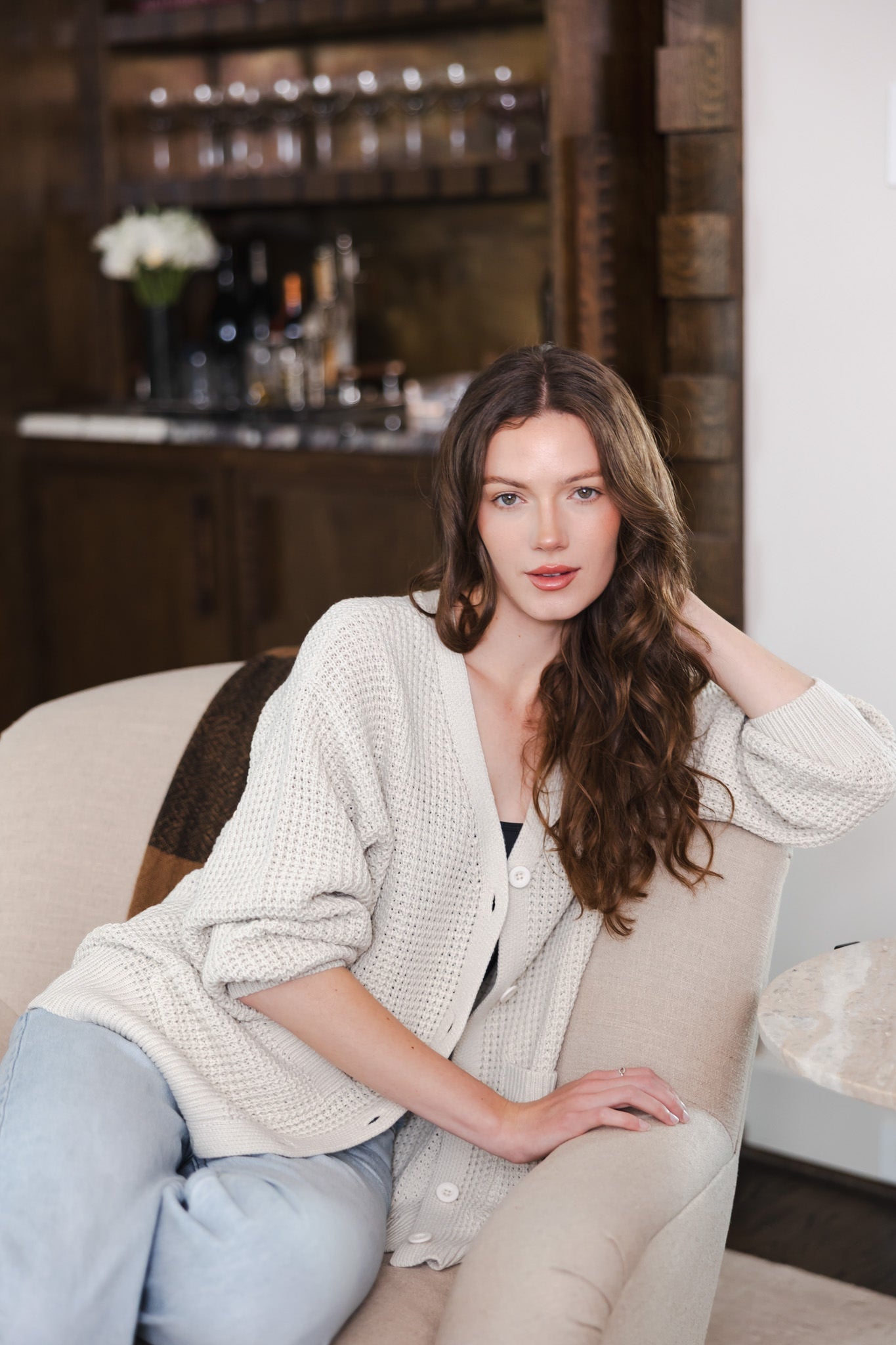 Modal sitting in a living room on a sofa wearing Chunky Knit Women's Waffle Cardigan in Oat over black camisole and blue jeans