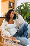 Woman sitting on a chair in a cozy room with a bookshelf and plant wearing a cream colored chunky knit cardigan, white camisole and jeans