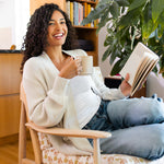 Woman wearing a chunky knit cardigan sitting in a chair reading a book and holding a mug in a cozy room.