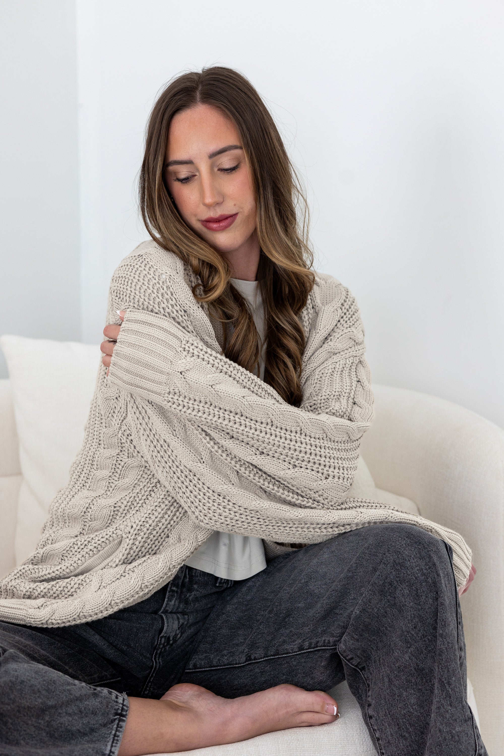 Woman sitting on a white couch snuggling in a oversized beige knitted cardigan.