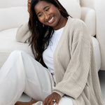 Woman sitting on a white couch wearing a beige cardigan and white pants.